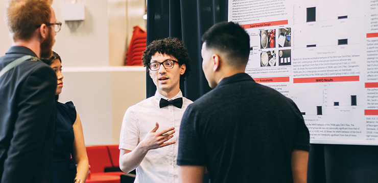 Student with bow tie presenting research to other students in front of poster