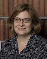 Photo of Fernanda, wearing a dark patterned shirt and glasses standing in front of book shelves