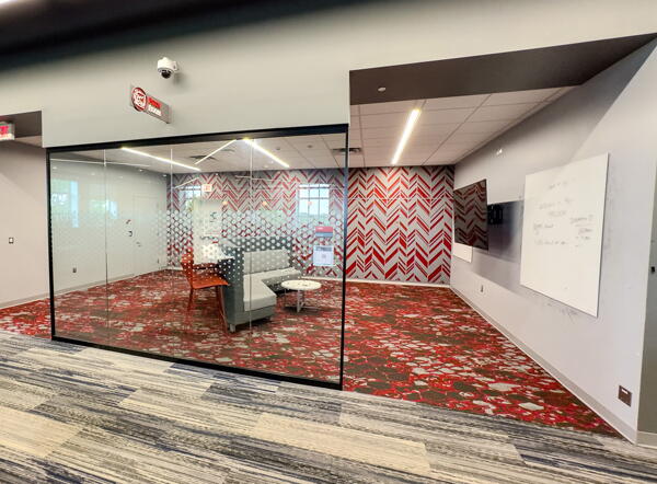 Glass wall looking into Pitch Room with seating and wall-mounted screen and whiteboards