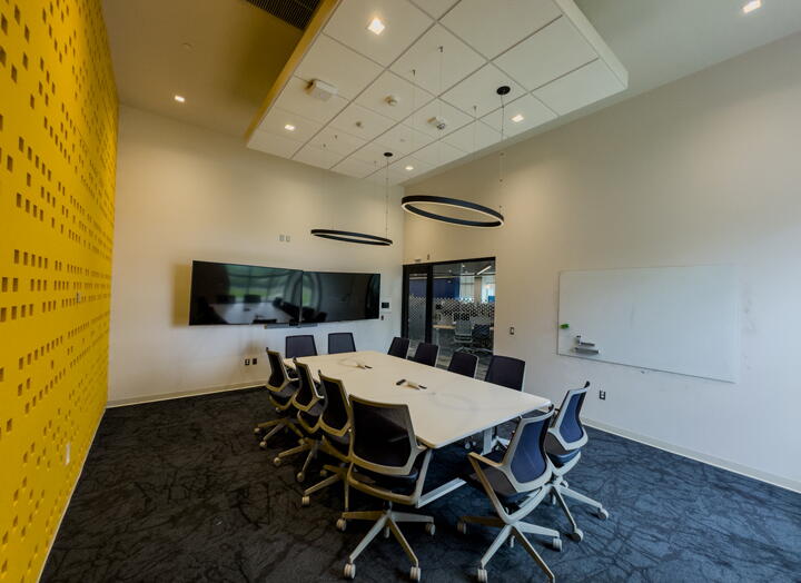 Interior of conference room with long table, 12 chairs, two displays, whiteboard and a yellow wall