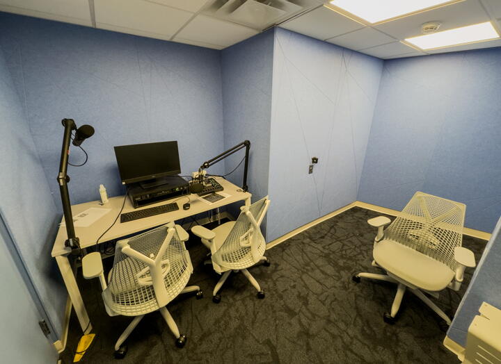 Audio booth, includes two chairs at an audio podcast station with a computer and microphones and another chair.