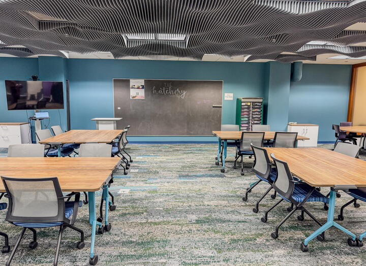 Classroom-style space with many group desks and chairs and a large chalkboard that says hatchery in cursive