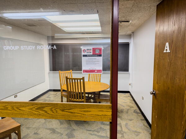 View through glass wall into group study room with table, chairs, and wall-mounted whiteboard and chalkboard