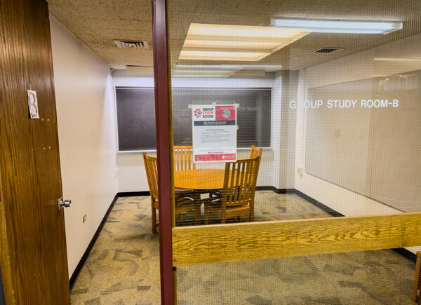 View through glass wall into group study room with table, chairs, and wall-mounted whiteboard and chalkboard