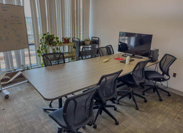 Conference room with large table with eight chairs, a rolling whiteboard, a monitor, plants, and a wall of windows with blinds.