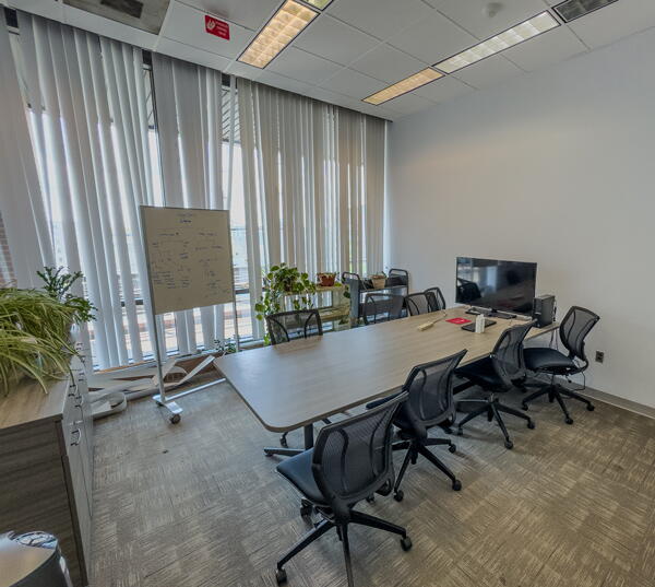Conference room with large table with eight chairs, a rolling whiteboard, a monitor, plants, and a wall of windows with blinds.